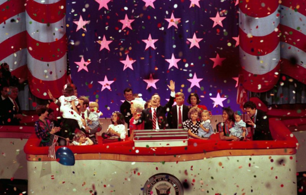 Eine Gruppe von Menschen auf einer Bühne mit einem Rednerpult, das ein Logo, Luftballons und Konfetti zeigt, vor einem Sternenhimmel und einem Vorhang.