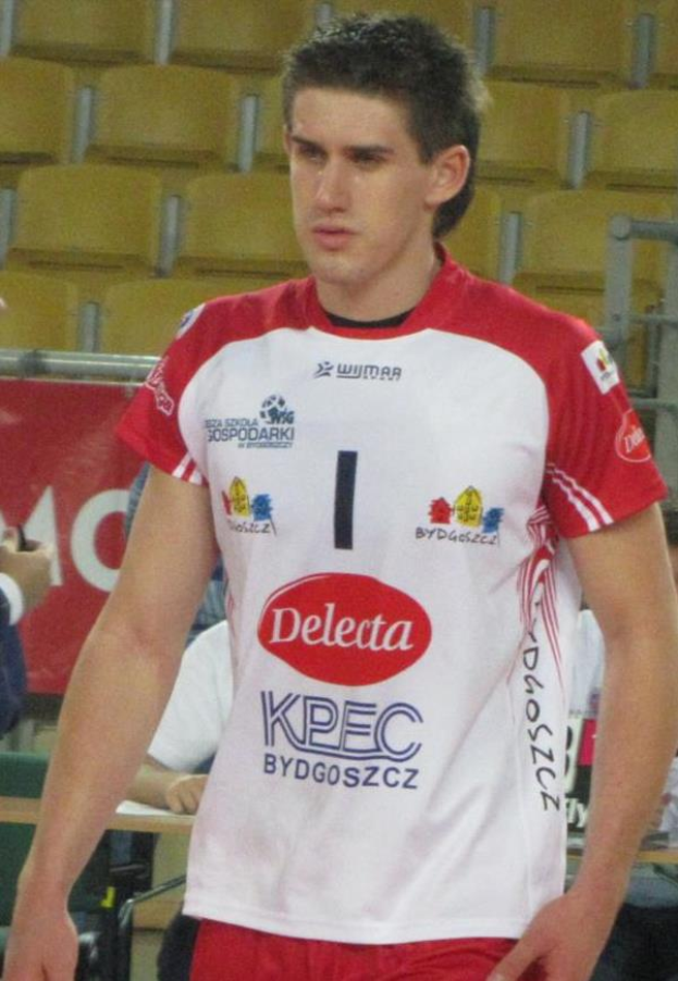 Ein Mann in einer roten und weißen Handballdressur steht auf einem Feld, entschlossen, mit Zuschauern und einer roten Fahne mit der Aufschrift "Krasnoyarsk vs Bydgoszcz" im Hintergrund.