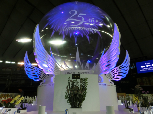 Ein großer Kristallball mit Flügeln oben drauf, umgeben von Blumenbouquets, Namensschildern und anderen Gegenständen auf einem festlichen Boden, mit Schildern, Lichtern und einem Bildschirm im Hintergrund.