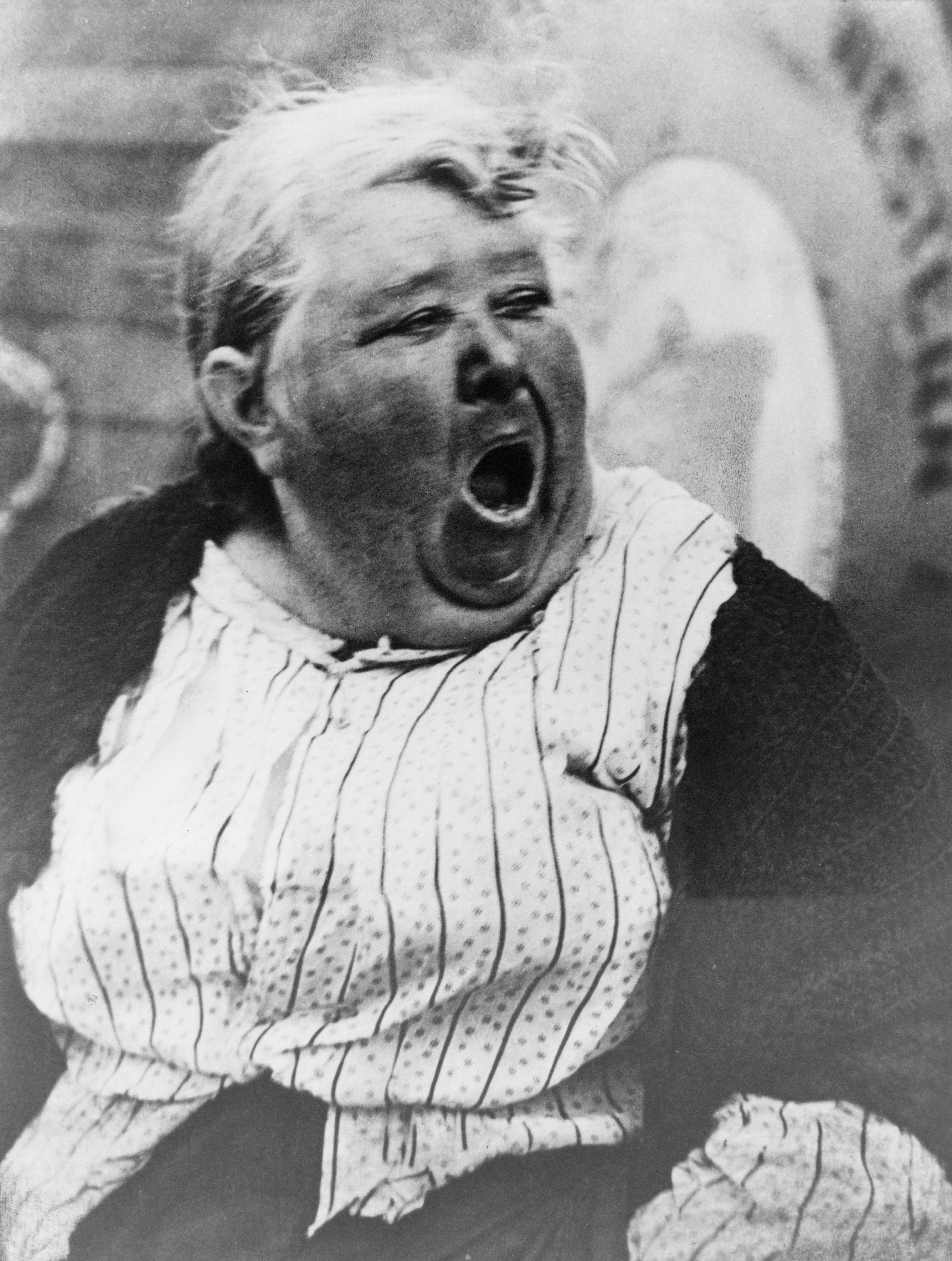 Ein Schwarz-Weiß-Foto einer Frau, die im Vordergrund schreit, mit einer Wand und einigen Gegenständen im Hintergrund.