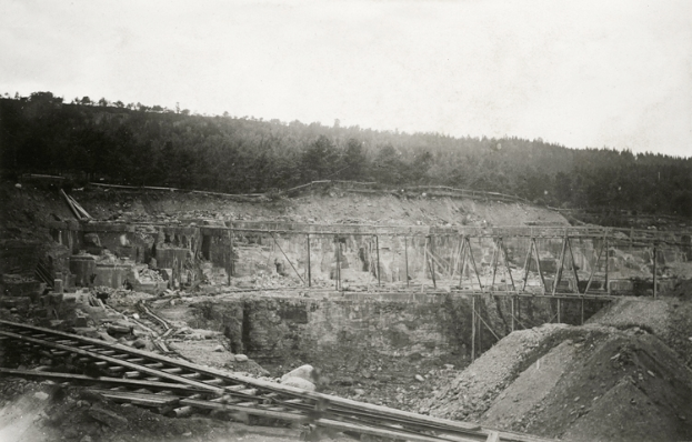 Ein Schwarz-Weiß-Foto einer Baustelle mit einer Brücke im Hintergrund, umgeben von Bäumen und einem klaren Himmel, mit Eisenbahnschienen und Steinen unten.