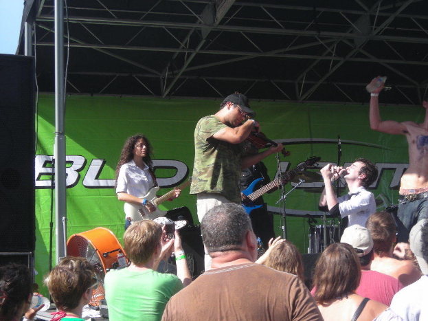 Eine Gruppe von Musikern, die verschiedene Instrumente auf einer Bühne spielen, während das Publikum davor steht und die Vorstellung genießt, mit einem großen schwarzen Lautsprecher links daneben und einer grünen Fahne im Hintergrund.