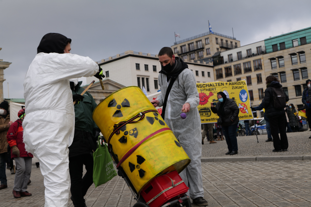 Eine Gruppe von Menschen in weißen Anzügen und Masken steht um einen gelben Eimer herum, mit einem Wagen im Vordergrund und Gebäuden, Fahnen und einem bewölkten Himmel im Hintergrund, einige halten Schilder mit Text.