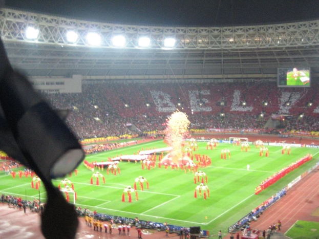 Ein Stadion mit Flutlicht, Eisengittern, Zuschauern, stehenden Personen, Werbetafeln, Anzeigetafeln, Überwachungskamera, Fahnen und Décor.