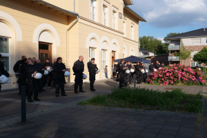 Gruppe von Polizisten vor einem Gebäude mit Fenstern und Türen stehend, Helme haltend, in der Nähe eines Zeltes, umgeben von Pflanzen, Blumen, Gras, einem Straßenschild, Bäumen und einem bewölkten Himmel.