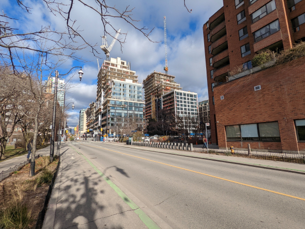 Eine Stadtstraße mit hohen Gebäuden, Straßenlaternen, Stromleitungen, Fahrzeugen, Fußgängern, Geländern, Bäumen und einem bewölkten Himmel.