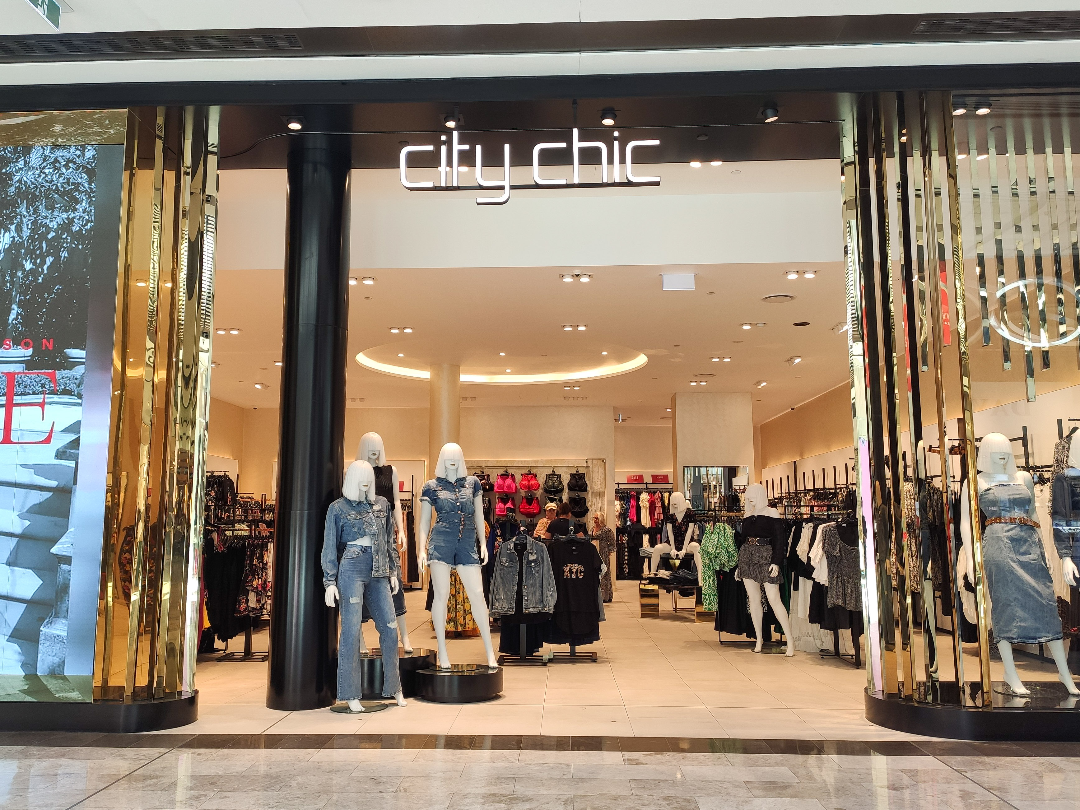 Ein stylischer Mode-Boutique in einem Einkaufszentrum mit Schaufensterpuppen in der neuesten Mode, Kleidung an Bügeln, einer Säule, einem Schild, Deckenleuchten und einem gefliesten Boden.