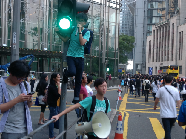 Ein Mann steht auf einer Ampel und hält ein Megafon und ein Handy in der Hand, umgeben von einer Gruppe von Menschen auf der Straße, mit Verkehrskegeln, einem Metallzaun, einem Schild, Fahrzeugen, Gebäuden, Bäumen und einem bewölkten Himmel im Hintergrund.