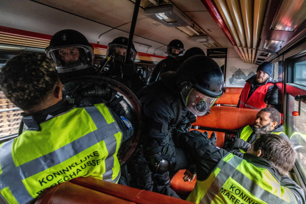 Polizeibeamte in Einsatzausrüstung auf einem Bus sitzend, mit einer Person in der Mitte und sichtbaren Fenstern auf der rechten Seite; ein Plakat ist im Hintergrund an der Wand zu sehen.