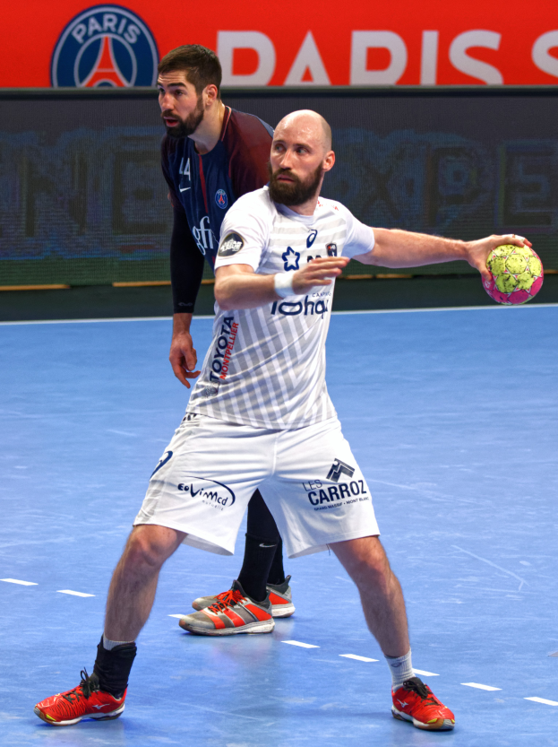 Zwei Männer spielen Handball auf einem Platz, einer hält einen Ball, mit einer Tafel im Hintergrund, auf der 'Paris Saint-Germain vs Paris Saint Germain' steht.