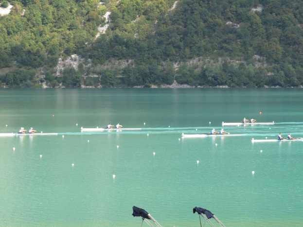 Eine Gruppe von Menschen bei einem Ruderwettbewerb auf einem Gewässer mit Bäumen und einem Hügel im Hintergrund, mit Kleidung über Stühlen am Boden.