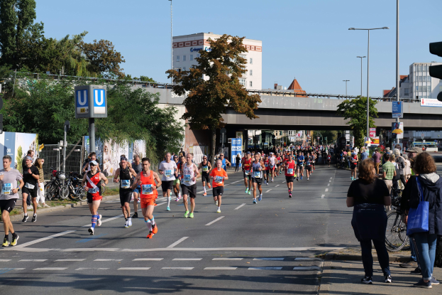 Eine Gruppe von Menschen, die bei einem Marathon auf einer Straße mit Bäumen, Laternenmasten und Schildern, Fahrrädern, einem Zaun, Gras, einer Brücke und Gebäuden läuft, unter einem klaren blauen Himmel.