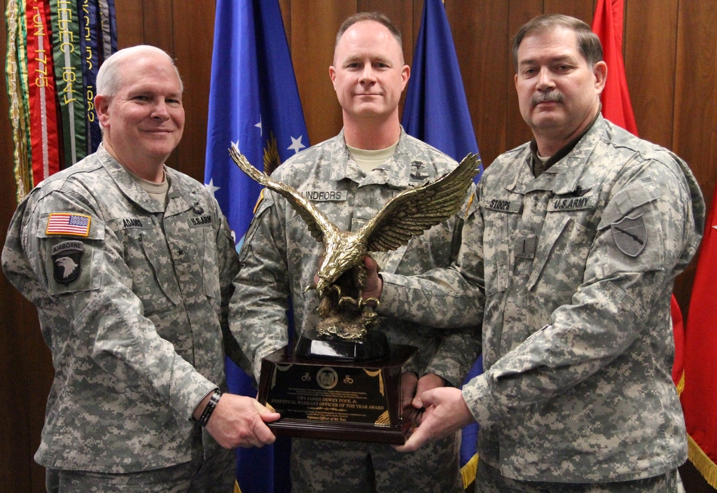 Drei Männer in Uniformen halten Auszeichnungen, mit Fahnen im Hintergrund und buntem Text in der oberen linken Ecke.