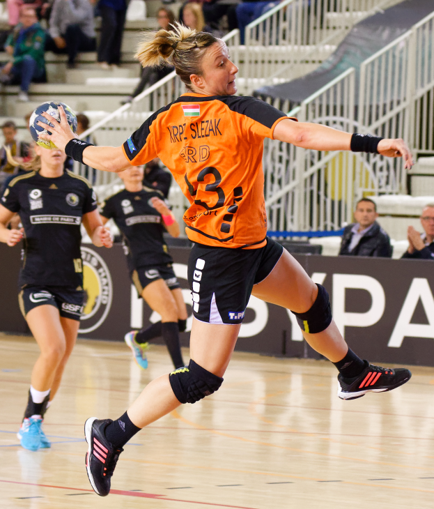 Eine Frau in einem orangen T-Shirt und schwarzen Shorts spielt Handball mit einem Ball in der Hand, umgeben von Menschen auf Treppen und Geländern und einem Banner mit der Aufschrift "Krp Slezak - Handball-Weltmeisterschaft 2019".