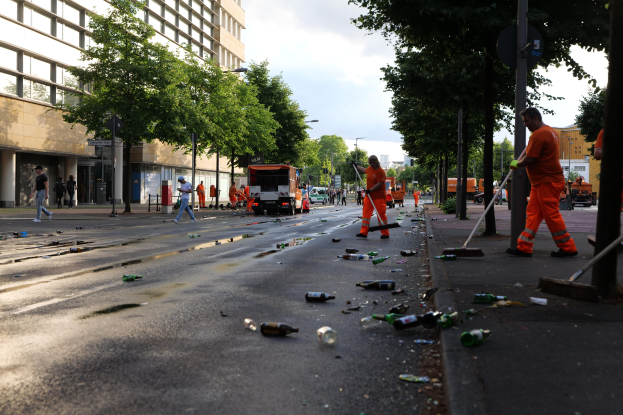 Eine Gruppe von Menschen in orangen Uniformen, die Müll auf einer Straße voller Flaschen und Schutt säubern, mit Bäumen, Pfählen und Fahrzeugen im Hintergrund unter einem bewölkten Himmel.
