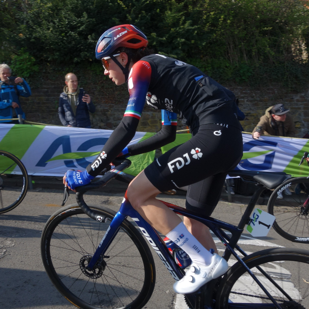 Eine professionelle Radrennfahrerin, Katie Archibald, fährt in Helm, Brille, Handschuhen und Schuhen ein Fahrrad eine Straße entlang an einer Menge vorbei, mit einem Banner, einer Wand, Bäumen und einem Gebäude im Hintergrund.