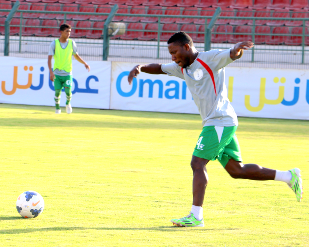 Ein Mann schießt einen Fußball auf einem grünen Feld in einem Stadion, mit zwei Personen in der Nähe und Bannern, Geländern und Stühlen im Hintergrund.
