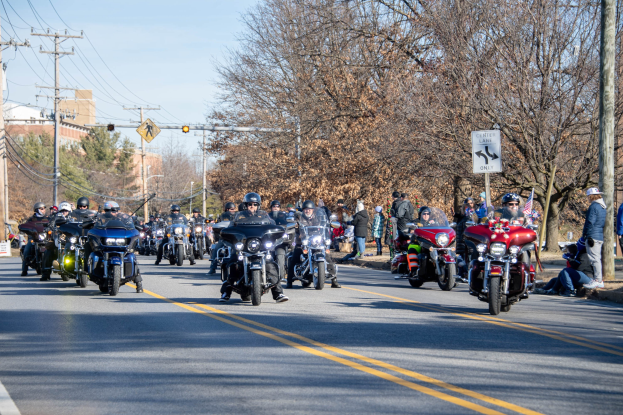 Eine Gruppe von Menschen auf Motorrädern fährt eine Straße entlang, die von Strommasten, Schildern, Bäumen und Gebäuden gesäumt ist, unter einem klaren blauen Himmel, wobei einige Fahrer Helme tragen.