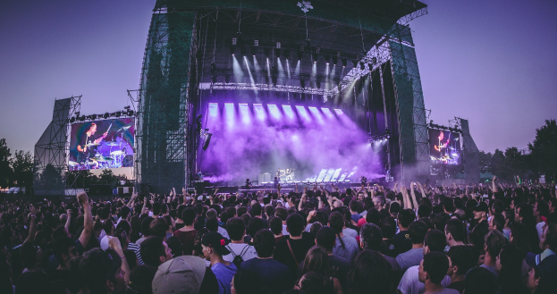 Eine große Menge Menschen auf einem Musikfestival mit einer Bühne, auf der Musiker spielen, beleuchtet von Lichtern und umgeben von Bildschirmen, vor einer Kulisse aus einem Gebäude, Bäumen und einem klaren blauen Himmel.