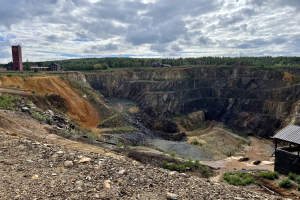 Großer Tagebau umgeben von Bäumen, Gebäuden und einem Schuppen in einem Schmutzfeld mit verstreuten Steinen und Pflanzen unter einem bewölkten Himmel.