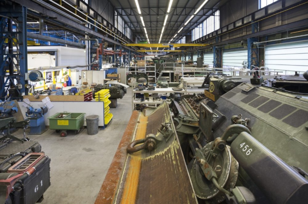 Ein großes Lagerhaus voller Maschinen, einschließlich eines Landfahrzeugs auf der rechten Seite, Metallobjekte auf der linken Seite und Lampen oben.