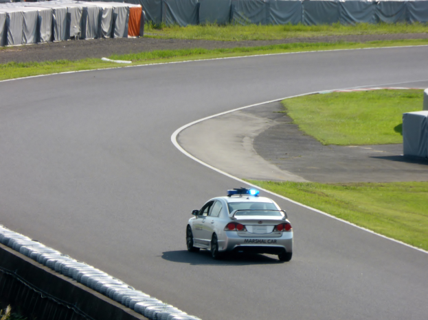 Ein Polizeiwagen fährt eine Rennstrecke entlang, mit Gras auf beiden Seiten und Menschen, Zelten und anderen Gegenständen im Hintergrund.
