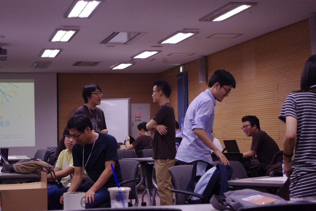 Ein Klassenzimmer mit Tischen, Stühlen, Laptops, Taschen, einer Projektionsfläche, einer Holzwand und einer Tür im Hintergrund.