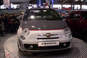 Ein Fiat 500 Abarth Auto auf einer Automesse ausgestellt, umgeben von Menschen und Bannern im Hintergrund.