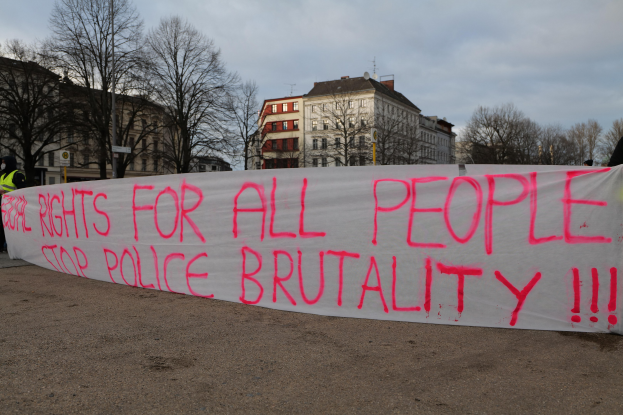 Gruppe von Menschen mit einem Banner mit der Aufschrift 'Rechte für alle Menschen Stoppt Polizeigewalt' vor einem Straßenschild, einem Schild, Bäumen, Gebäuden und einem bewölkten Himmel.