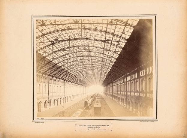 Schwarz-weiß-Foto eines Zuges an einem Bahnhof mit Gebäuden, metallgedecktem Bahnsteig und Text unten.