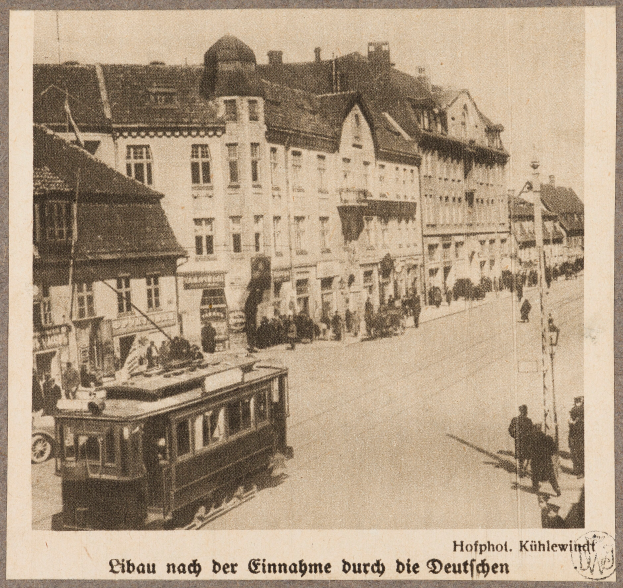Ein altes Schwarz-Weiß-Foto einer Stadtstraße mit einer Tram, Gebäuden, Straßenlaternen, Straßenlaternen und Passanten, mit Text am unteren Rand.