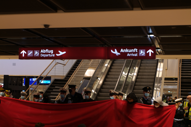 Eine Gruppe von Menschen mit M├╝tzen und Masken steht vor einer Rolltreppe am Flughafen, h├Ąlt ein rotes Tuch und positioniert sich unter Schildern mit Text, wahrscheinlich beim Einsteigen in ein Flugzeug w├Ąhrend der Coronavirus-Pandemie.
