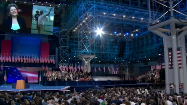 Eine große Menschenmenge sitzt vor einer Bühne mit einem Rednerpult, Mikrofon, Flaggen, einem Bildschirm und Beleuchtungsausrüstung und wartet auf eine Präsidentschaftsdebatte.