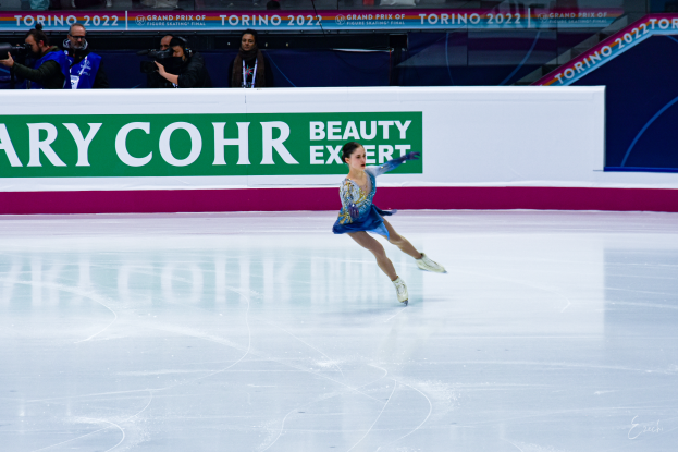 Eine Frau in einem blauen Kleid, die auf einer Eisfläche Schlittschuh läuft, umgeben von Zuschauern mit Kameras, mit einem Schild im Hintergrund, auf dem 'Tessa Virtue und Scott Moir' steht.