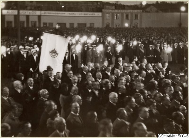 Schwarzes und weißes Foto einer großen Menge, die vor einem Gebäude steht, mit einer Person, die eine Flagge hält, und Polen und Lichter im Hintergrund sowie dem Text "Gustav Klimts Rede an der Berliner Mauer" unten.