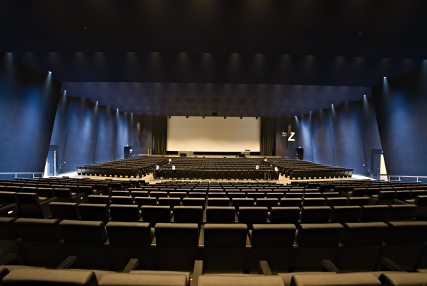 Ein großer Zuschauerraum mit Reihen von Stühlen, einem Projektionsschirm und Bühnenobjekten, beleuchtet von Wandlampen.