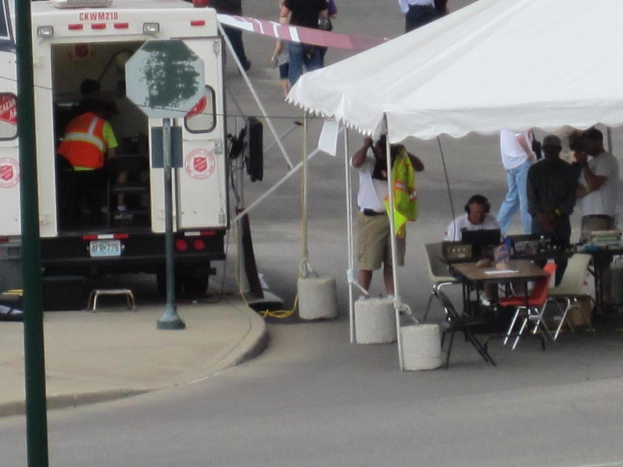 Ein Rettungswagen steht links von einer Straße, während rechts ein Zelt eine Gruppe von Menschen schützt, bei der ein Tisch und ein Stuhl in der Nähe stehen.