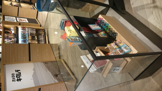 Kartons, Flaschen, Bücher und Poster mit Text sind auf Glas-Tischen und Holzregalen angeordnet.