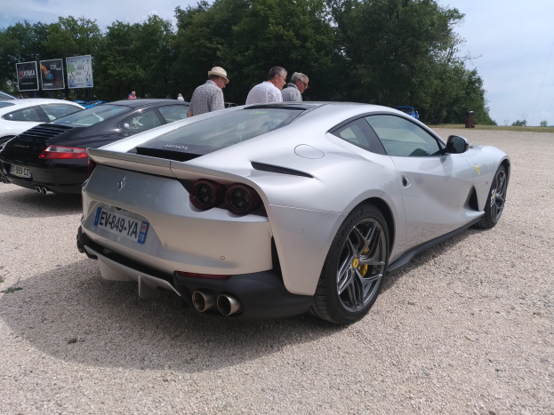 Roter Ferrari 812 Superfast auf dem Goodwood Festival of Speed mit Menschen in Kappen, Bäumen, Texttafeln und bewölktem Himmel im Hintergrund.