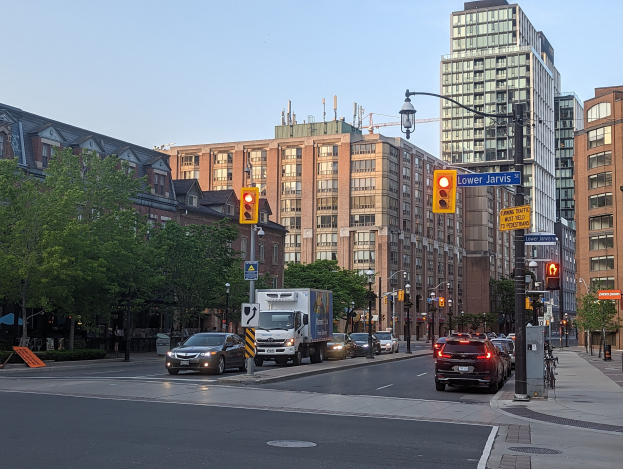 Stadtstraße mit starkem Verkehr, einschließlich eines Lastwagens, eines Autos und eines SUV, umgeben von Laternenmasten, Ampeln, Texttafeln, Bäumen und Gebäuden vor einem Himmel als Hintergrund.