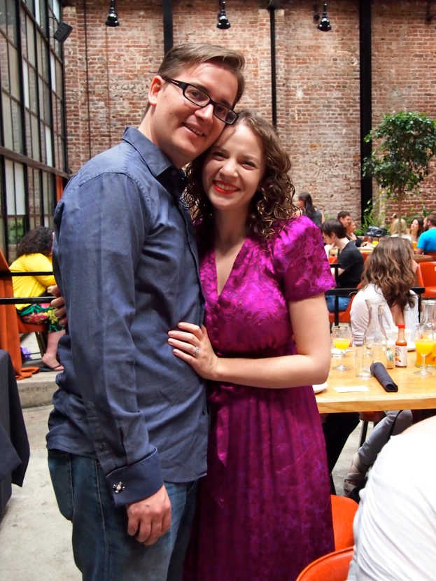 Ein Mann und eine Frau lächeln vor einer Backsteinwand mit Lichtern, während Menschen an Tischen mit Gläsern, Flaschen, Säften und einer Pflanze im Hintergrund sitzen.