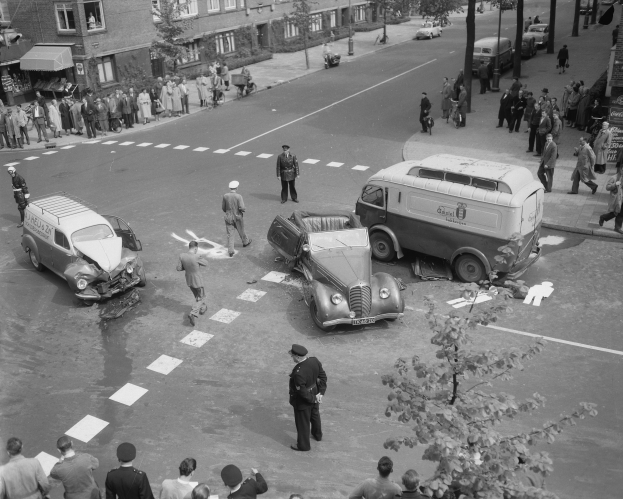 Schwarz-weißes Bild eines Autounfalls an einer Straßenkreuzung mit einem Van, mehreren Fahrzeugen und einer Gruppe von Menschen in der Nähe, vor dem Hintergrund von Gebäuden, Bäumen, Pfählen und Texttafeln.