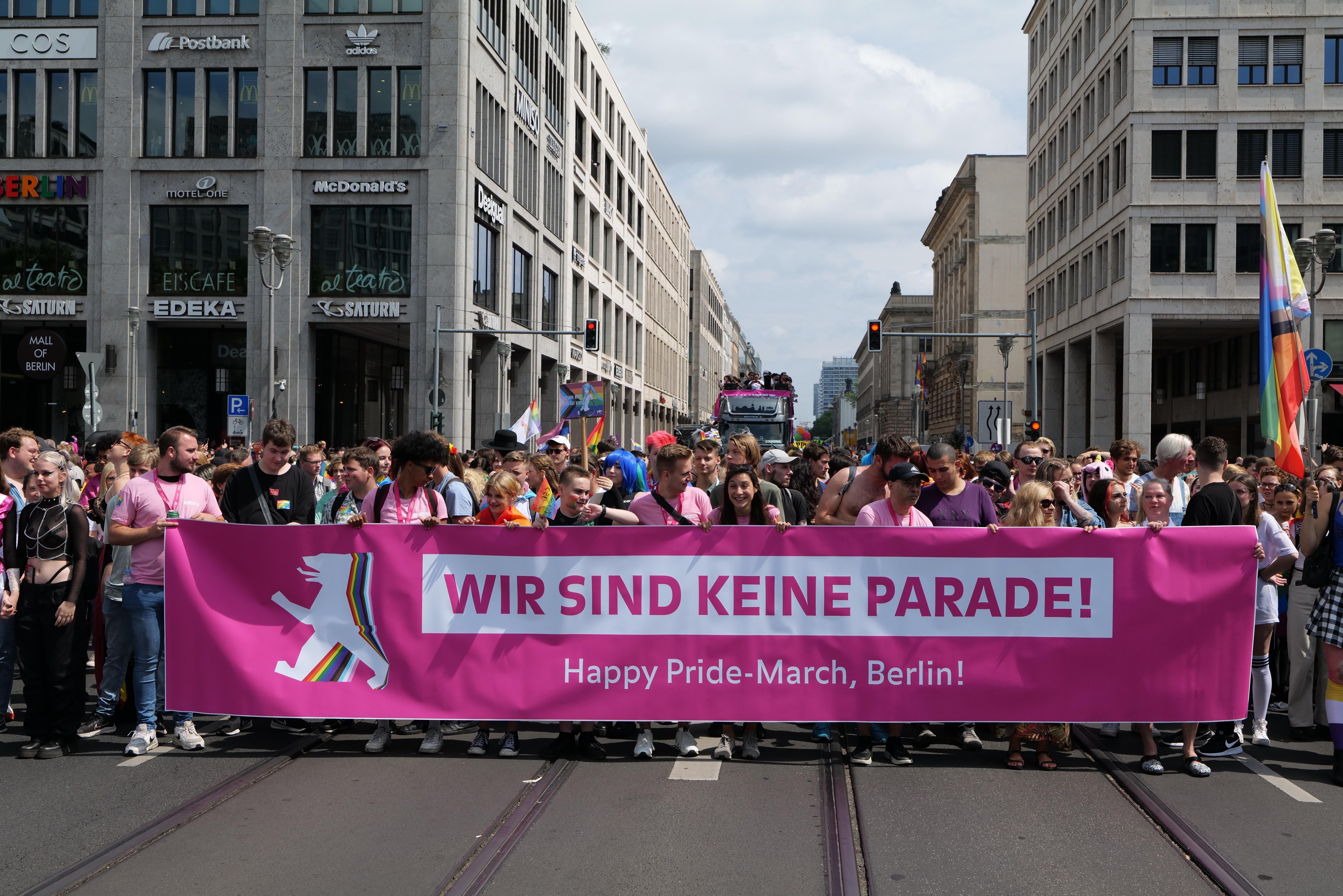 Eine Gruppe von Menschen marschiert auf einer Straße in Berlin, Deutschland, mit einem pinken Banner mit der Aufschrift "Happy Pride March", mit Gebäuden, Laternenmasten und Verkehrszeichen an der Straße.