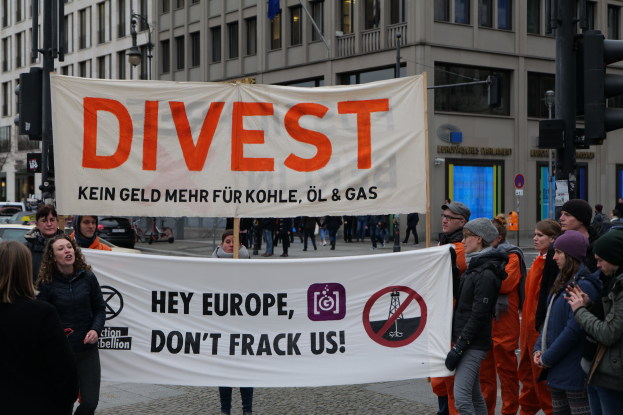 Eine Gruppe von Menschen steht auf der Straße und hält ein Banner mit der Aufschrift "Divest Hey Europe, Don't Frack Us", mit Gebäuden, Straßenlaternen, Fahrzeugen, Bäumen und einem klaren blauen Himmel im Hintergrund.