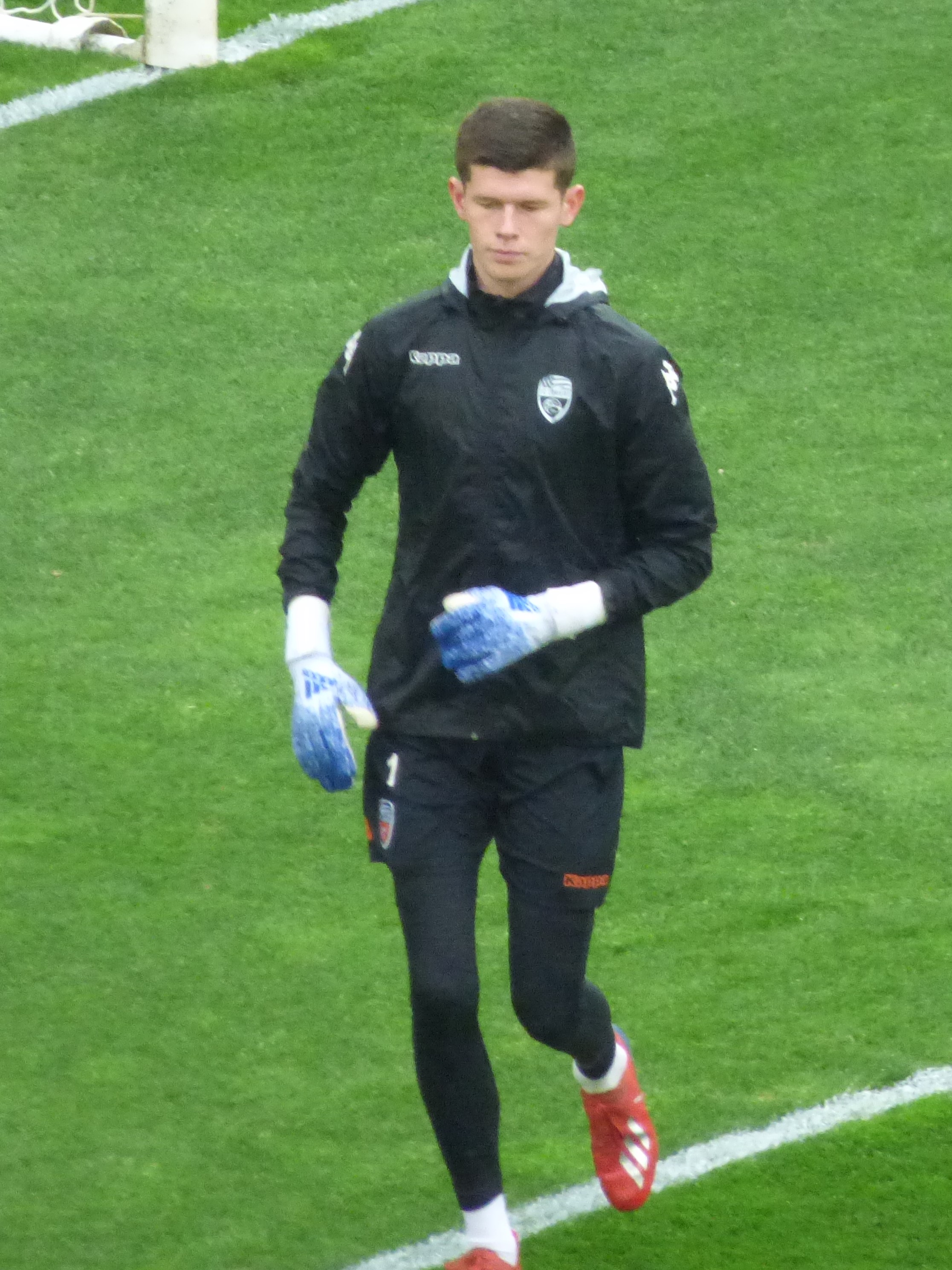 Ein Mann in einer schwarzen Jacke und blauen Handschuhen geht auf einem Fußballfeld mit einem Tor im Hintergrund, trägt Fußballhandschuhe.