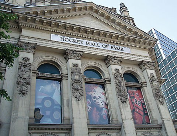 Außenansicht des Hockey Hall of Fame in New York City mit Glasfenstern, lesbarem Text an der Fassade, einem Baum auf der linken Seite und einem Teil des Himmels darüber.