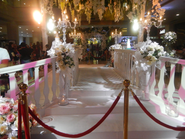 Eine Trauung in einem Einkaufszentrum mit weißem Teppich, Geländern, Blumenschmuck und Deckenbeleuchtung, bei der viele Menschen anwesend sind.