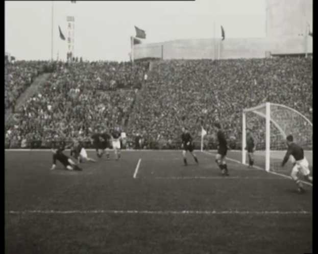 Ein Schwarz-Weiß-Foto eines WM-Finalspiels 1958 zwischen Manchester United und Liverpool mit Spielern auf dem Feld, einem Torpfosten auf der rechten Seite, Zuschauern in den Rängen und Fahnen im Hintergrund.