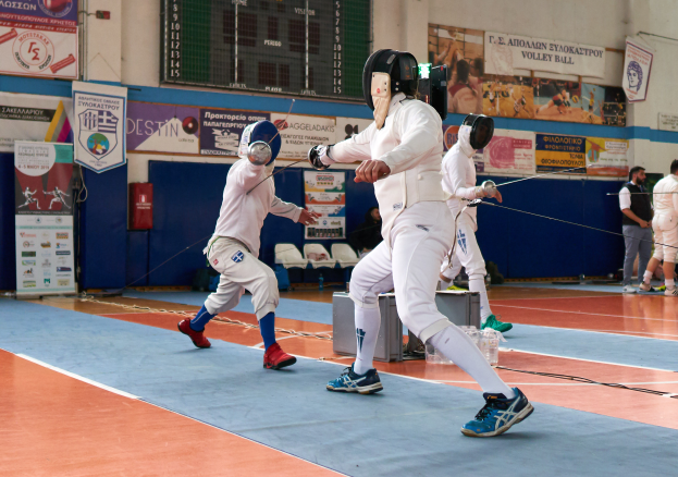Eine Gruppe von Menschen in Helmen und mit Schwertern ficht in einer Turnhalle mit Bannern, einem Bildschirm, Stühlen und anderen Gegenständen herum.