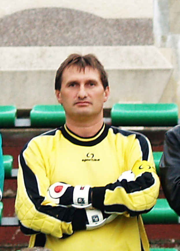 Ein Mann in einem gelben Trikot, wahrscheinlich ein Torwart, steht neben einem anderen Mann in einer schwarzen Jacke, mit grünen Stühlen und einer Wand im Hintergrund.